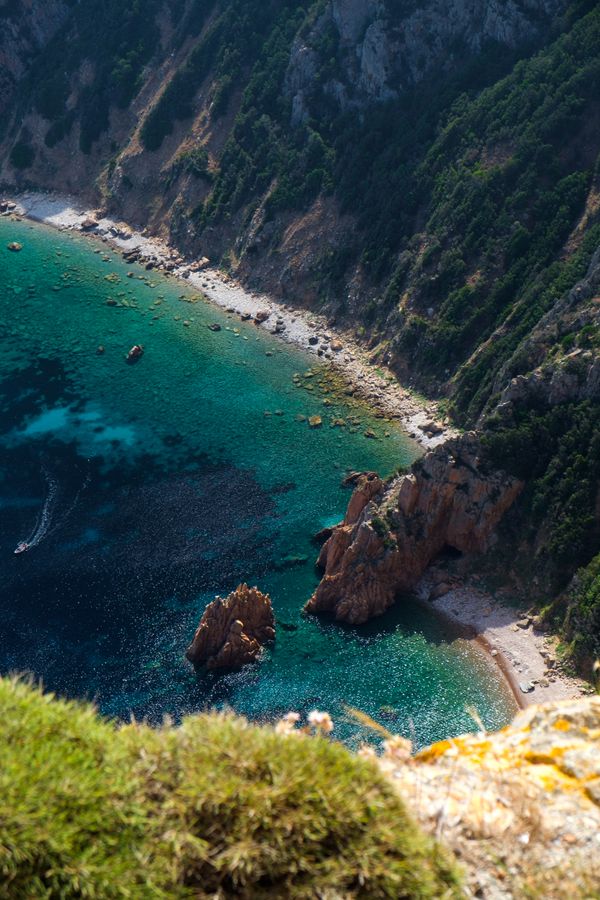 Naar beneden kijkend vanaf de Capo Rosso is een azuurblauwe zee in de diepte te zien, in het midden van het beeld, omringt door een kleine rand strand, steile rotswanden ver omhoog. Aan de rand van het beeld een kleine boot.