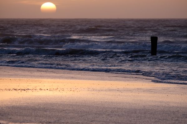 De ondergaande zon boven de zee, golven slaan stuk op het strand. Rechts staal een paal in het water. Het licht reflecteert in het strand.